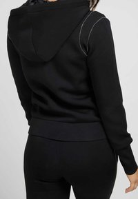 Svart huvtröja i en smidig, matt textur, med vit sömnad längs axelsömmarna. Tätsittande design med ribbat avslut.