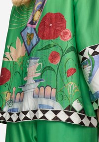 Tessuto verde con motivi floreali vivaci e artistici, che presenta una fontana e un bordo decorativo con motivi geometrici in bianco e nero.