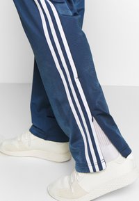 Námořnické sportovní kalhoty s bílými postranními pruhy, vybavené zipy na kotnících. Vyrobené z hladkého materiálu, nošené s bílými teniskami.