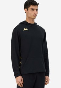 Felpa nera con cappuccio e logo dorato sul petto a sinistra, arricchita con dettagli dorati lungo le maniche. Realizzata in un tessuto morbido e strutturato. Include una tasca frontale.