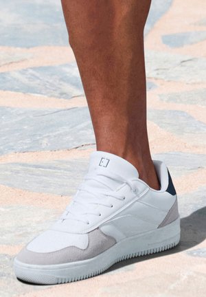 Weißer Sneaker mit grauen Wildlederakzenten und dunkelblauem Absatz. Schnürdesign, strukturierte Gummisohle und gepolsterter Kragen für Komfort.