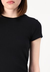 Top scurt negru cu mânecă scurtă și decolteu rotund, având un design ajustat și o textură netedă, evidențiind elasticitatea materialului.