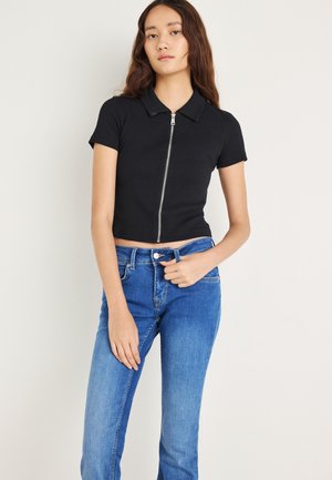 Top tipo polo negro recortado con cremallera frontal, mangas cortas y textura acanalada, combinado con jeans de talle alto en azul claro.