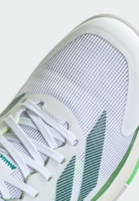 White athletic shoe featuring a mesh upper with green accents, textured design, and flat laces. Sole is cushioned with a rubber tread.