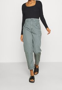 Top a maniche lunghe nera a coste abbinato a pantaloni larghi di colore verde pallido con tasche laterali e chiusura a bottone, indossato con sandali neri scivolati.
