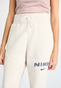 Světlé béžové tepláky s vysokým pasem, elastickým stahováním a vyšitým navy modrým logem "NIKE" na levé noze. Měkká textura.