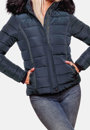 Donna con giacca trapuntata nera con cappuccio in pelliccia e mani nelle tasche, abbinata a jeans blu in denim.