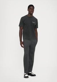 Man wearing dark gray t-shirt, gray dress pants, and black-and-white loafers standing against a plain light background.