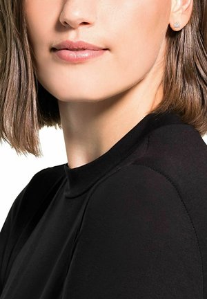 Primer plano de una persona que lleva una camiseta negra ajustada con un cuello alto. Se pueden ver pendientes de plata con forma de pulgar, y su cabello corto y castaño enmarca el rostro.