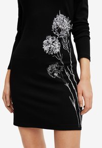 Svart klänning med vit blomsterbroderi på ena sidan, långa ärmar, figurnära passform och mjukt tyger.