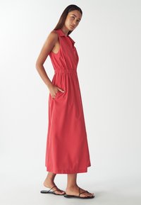 Czerwona, bezrękawnikowa sukienka maxi z kołnierzykiem, elastycznym paskiem w talii i bocznymi kieszeniami. Stylizowana z czarnymi sandałami na ramiączkach i gładką fakturą.