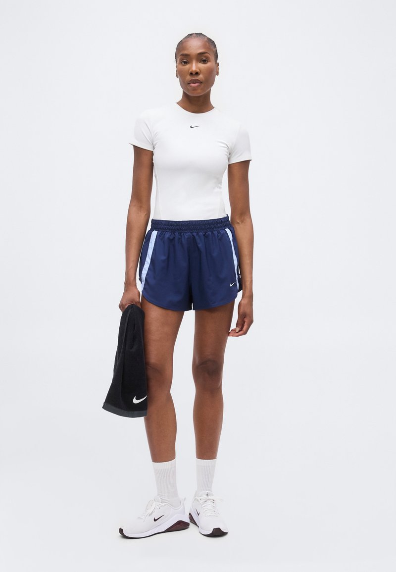 Mujer de pie de frente, vistiendo una camiseta blanca ajustada de manga corta, pantalones cortos deportivos azul marino, zapatillas blancas, sosteniendo una toalla negra en la mano derecha.