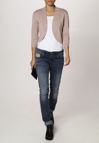 Ljusrosa kort kofta med trekvartsärmar över en vit topp, kombinerad med mörkblå jeans och svarta ankelboots.