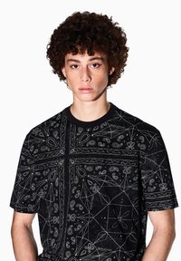 T-shirt nera a maniche corte con intricati motivi paisley e geometrici bianchi, scollatura rotonda e texture morbida del tessuto.