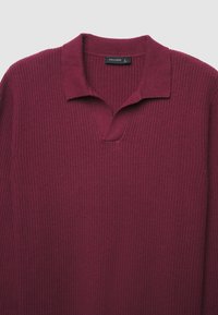 Maglione bordeaux a costine con colletto piatto e scollatura a V. Presenta un tessuto testurizzato e un design minimalista. Etichetta del marchio visibile.