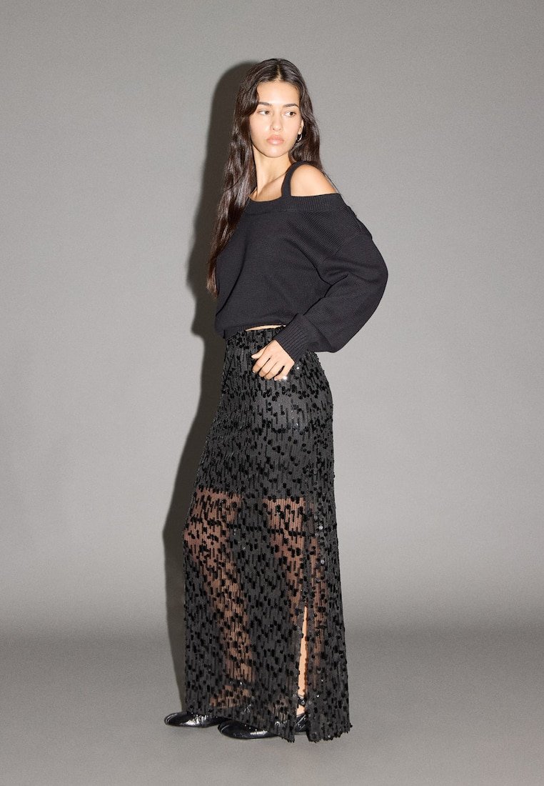 Maglione nero a costine con design scivolato sulle spalle abbinato a una gonna lunga nera trasparente con motivi intricati simili a perline e spacchi.
