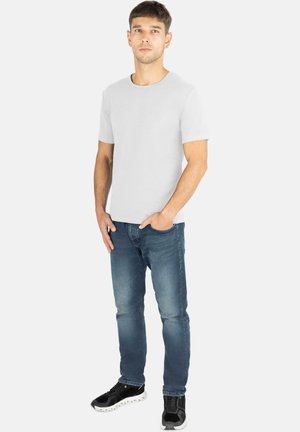 Weißes T-Shirt mit kurzen Ärmeln und Rundhalsausschnitt, kombiniert mit blauen Jeans und schwarzen Sneakers mit strukturiertem Profil, stehend vor einem einfarbigen Hintergrund.