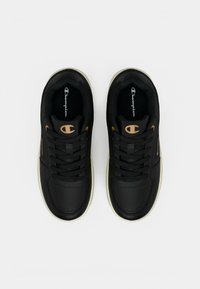 Ζευγάρι μαύρα αθλητικά παπούτσια Champion με χρυσό λογότυπο στη γλώσσα και στο πλάι, μαύρα κορδόνια και λευκές σόλες, από ψηλά σε λευκό φόντο.