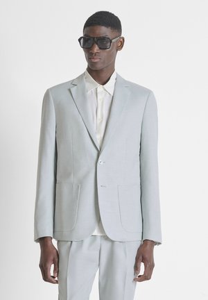 Muž v světle šedém obleku, bílé košili s knoflíky a velkých černých slunečních brýlích na jednobarevném bílém pozadí.