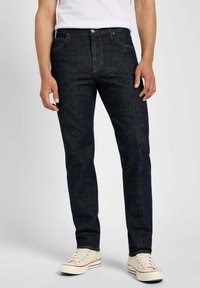 Jeans en denim indigo foncé avec une coupe slim, dotés de cinq poches, d'une fermeture éclair et d'un bouton en laiton. Portés avec des baskets de couleur claire.