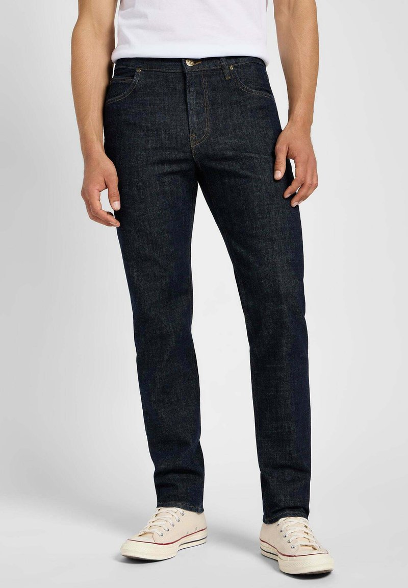 Jeans en denim indigo foncé avec une coupe slim, dotés de cinq poches, d'une fermeture éclair et d'un bouton en laiton. Portés avec des baskets de couleur claire.