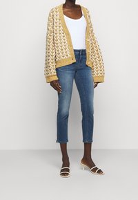 Cárdigan de punto mostaza con patrón de zigzag blanco, camiseta sin mangas blanca, jeans cortos azules y sandalias de tacón blancas. Textura suave, corte casual.