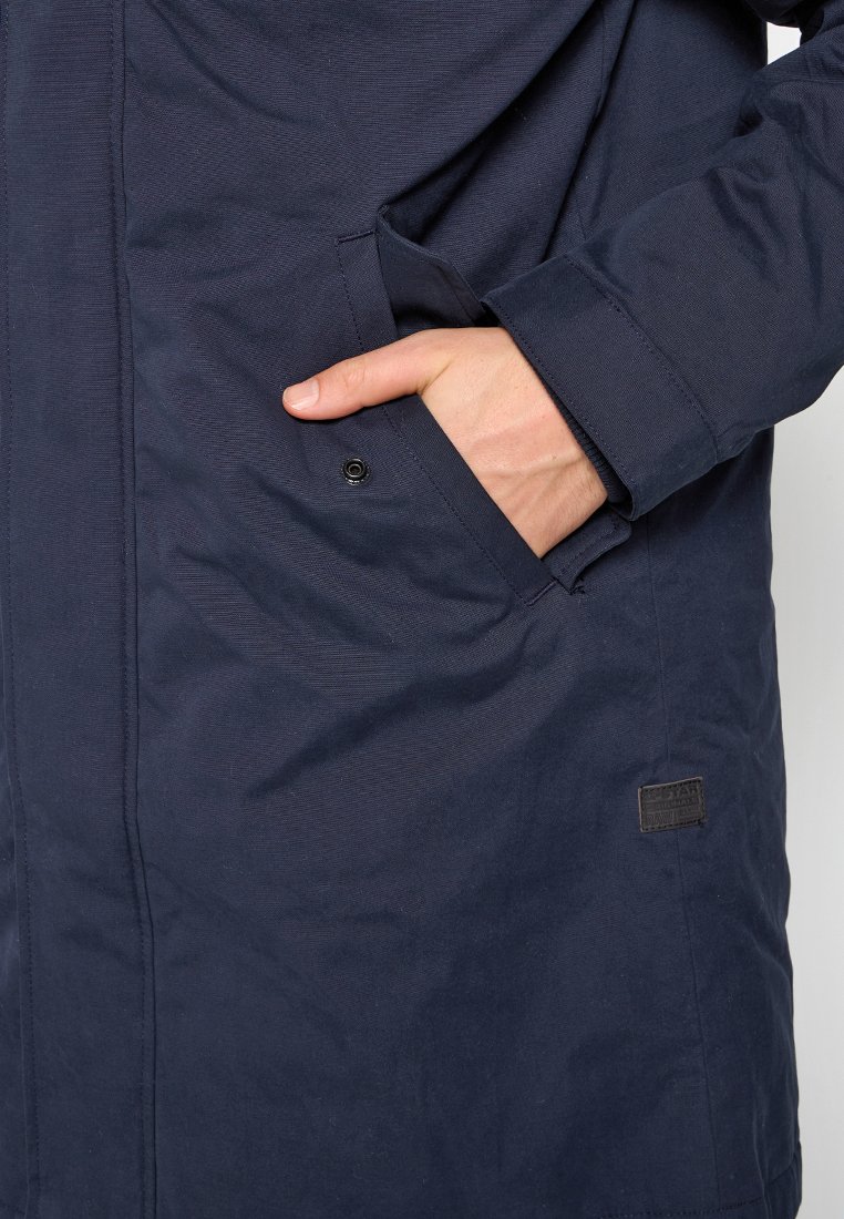 Main partiellement dans la poche d'une veste bleu marine avec des coutures visibles et une petite patch noire près de la poche.