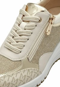 Sneaker beige con una toma in tessuto dorato testurizzato, una cerniera laterale e dettagli metallici. I lacci sono spessi e intrecciati.
