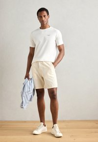 Witte t-shirt met korte mouwen, lichtbeige shorts, een blauw gestreept shirt vasthoudend en lichte sneakers dragend. Gladde texturen en casual ontwerp.