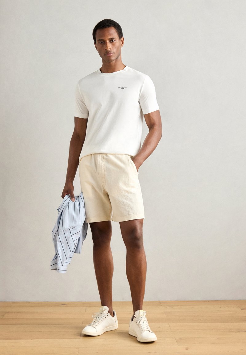 Witte t-shirt met korte mouwen, lichtbeige shorts, een blauw gestreept shirt vasthoudend en lichte sneakers dragend. Gladde texturen en casual ontwerp.