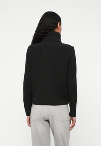 Črna rebrasta pletena turtleneck pulover z dolgimi rokavi. Ima visok ovratnik in skrajšan dizajn, v kombinaciji z svetlo sivo hlačami.