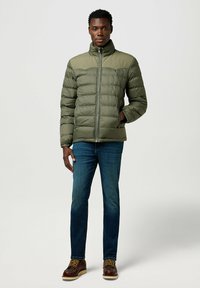 Olivgrön puffjacka med hög krage, dragkedjestängning och quiltat mönster. Kombinerad med blå jeans och bruna snörkängor.