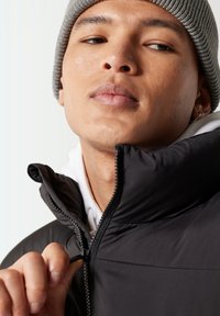 Chaqueta acolchada negra con un cuello alto, con cierre de cremallera. La persona lleva un gorro de punto gris y una sudadera blanca debajo.