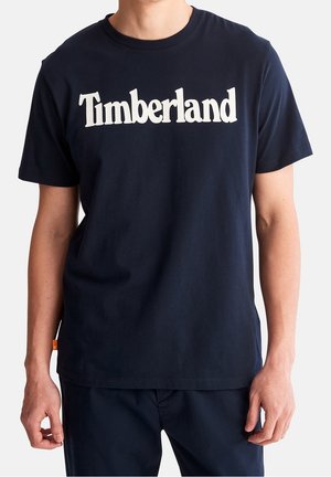 Mężczyzna ubrany w granatowy t-shirt z białym napisem "Timberland" na piersi, w parze z granatowymi spodniami na jednolitym tle.