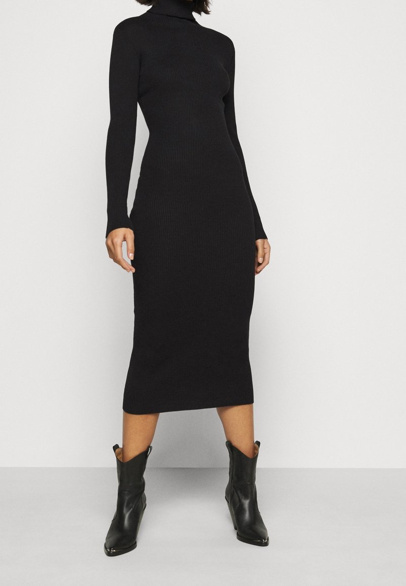 Robe noire côtelée à col roulé, manches longues, silhouette ajustée, longueur au genou. Portée avec des bottines noires pointues au design western.