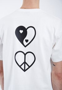 Camiseta blanca de algodón con dos grandes diseños de corazones negros en la parte posterior, uno con corazones más pequeños y otro con un símbolo de paz en su interior.