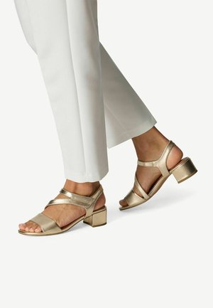 Guld læderåbne sandaler med krydsremme og blokhæl, kombineret med hvide skræddersyede bukser, der fremviser et slankt design og en glat tekstur.