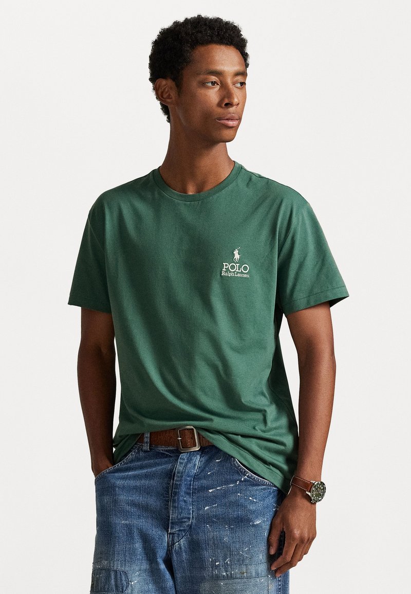Green cotton t-shirt with a crew neck, featuring a white embroidered logo on the left chest. Paired with blue denim shorts.