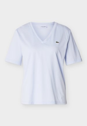 Chemise bleu clair à col en V et manches courtes avec un petit logo crocodile vert sur le côté gauche de la poitrine, présentée sur un fond uni.