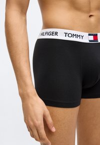Οι μαύροι μπόξερ έχουν λευκή μέση με το λογότυπο "TOMMY HILFIGER". Το ύφασμα φαίνεται λείο με εφαρμοστή σχεδίαση, αναδεικνύοντας το σχήμα.