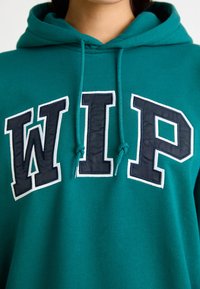Felpa con cappuccio color teal, caratterizzata da ricamo "WIP" in navy con dettagli bianchi, cappuccio con lacci e tessuto morbido e texture.