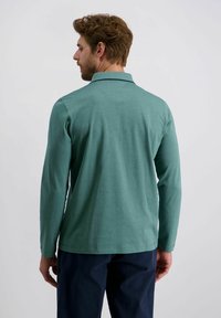 Mann mit Bart und lockigem Haar, von hinten gesehen, trägt ein langärmeliges türkisfarbenes Poloshirt und dunkelblaue Hosen vor einem einfarbigen Hintergrund.