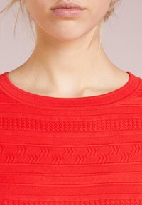 Gros plan sur le bas du visage et le cou d'une personne portant un pull en tricot texturé rouge vif avec un col rond.