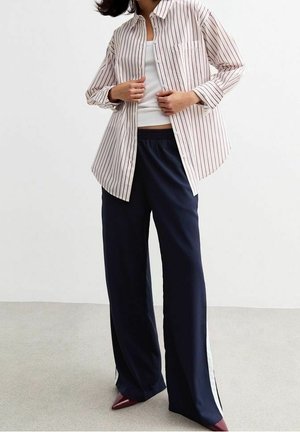 Kobieta ubrana w biały top na ramiączkach, białą koszulę na guziki w czerwone paski, granatowe spodnie o szerokich nogawkach z białymi paskami po bokach oraz bordo buty na szpic.