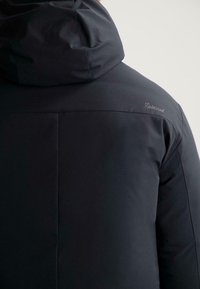 Černá zateplená bunda s kapucí, vyrobená z hladkého materiálu. Má prošité detaily a decentní logo na rameni.