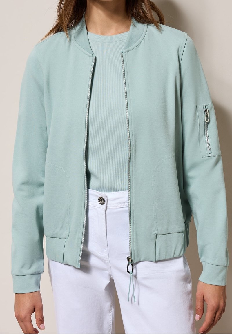 Kobieta w jasnoniebieskiej bomberce zapinanej na zamek, nosząca pod nią prążkowany jasnoniebieski top oraz białe spodnie, na beżowym tle.