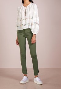 Blusa blanca con bordado floral y mangas abullonadas, combinada con pantalones ajustados verdes con dobladillos sin rematar y zapatillas blancas.