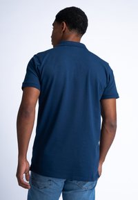 Tmavě modré polo tričko s krátkým rukávem, límečkem a texturovanou tkaninou. Volný střih, viditelné švy a rovný lem. Nosené s džíny.