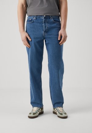 Person iført blå straight-leg jeans, grå skjorte og grønne, hvide og brune sneakers, der står mod en ensfarvet baggrund.