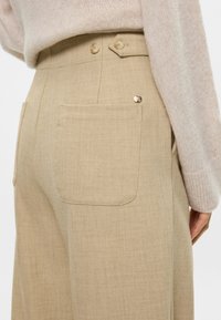 Beige ψηλόμεσες παντελόνες από υφασμάτινη ύφανση, με δύο πίσω τσέπες και δύο κουμπιά κοντά στη μέση.